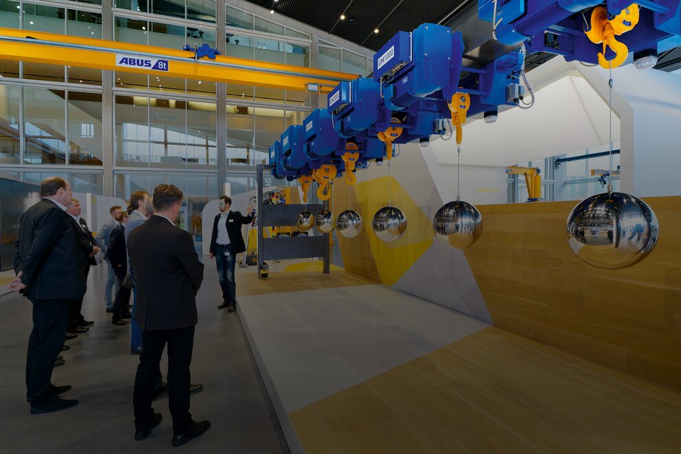 A group of visitors in the crane house is being shown how wire rope hoists work. The visitors are wearing formal clothing. At the top in the background is a ZLK overhead travelling crane with a load capacity of 8 tonnes.