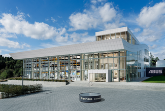Exterior view of the ABUS ‘KranHaus’ exhibition centre. Various types of crane systems can be seen behind the glass façade. In the foreground is an illuminated, round object with the address: KranHaus Archimedesstraße.