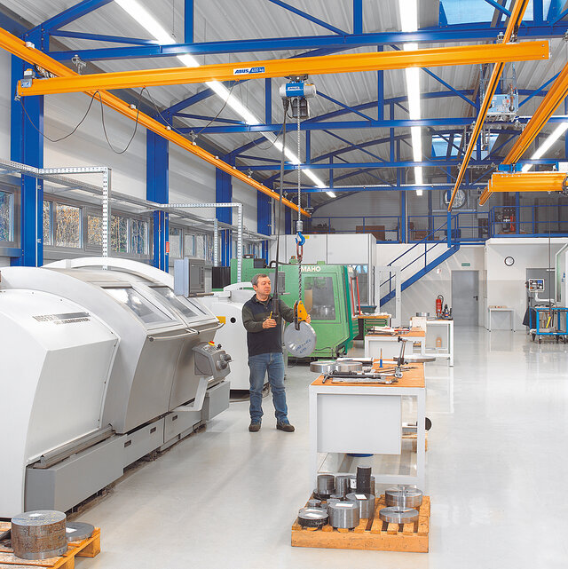 An employee operates a suspended rail system with a chain hoist and moves a load around a workshop