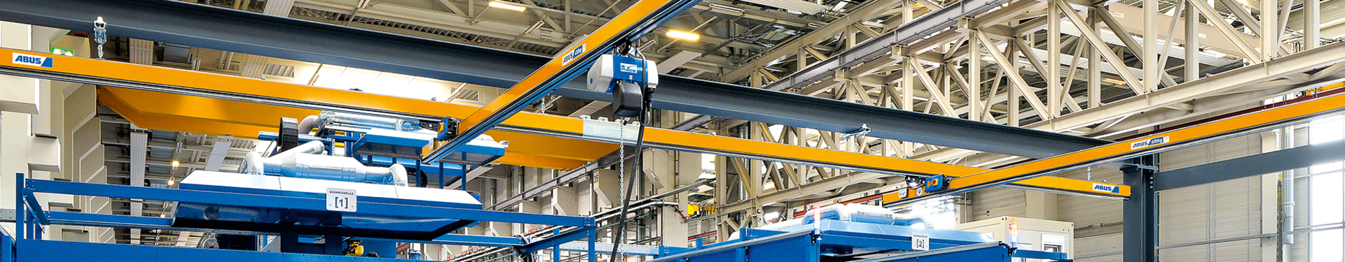 Several single-girder underslung overhead cranes of type EHB can be seen in an industrial hall. In the foreground, a worker operates a chain hoist with a pendant control. The chain hoist runs on a trolley in the profile of the main girder.