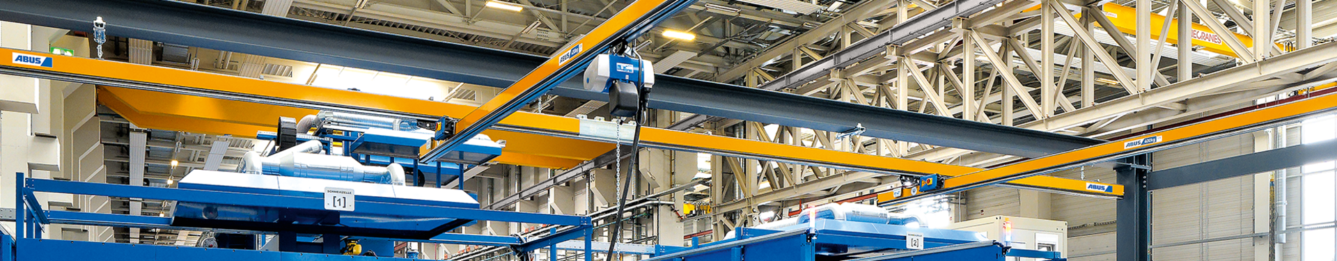 Single-girder underslung overhead crane type EHB Several single-girder underslung overhead cranes of type EHB can be seen in an industrial hall. In the foreground, a worker operates a chain hoist with a pendant control. The chain hoist runs on a trolley in the profile of the main girder.