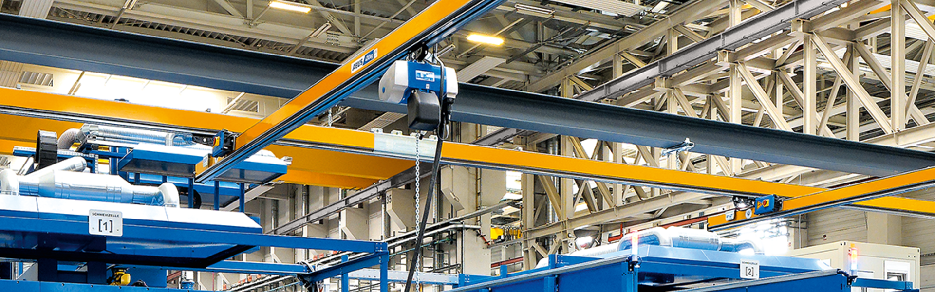 Several single-girder underslung overhead cranes of type EHB can be seen in an industrial hall. In the foreground, a worker operates a chain hoist with a pendant control. The chain hoist runs on a trolley in the profile of the main girder.