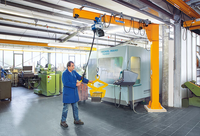 An employee is operating a column-mounted jib crane and lifting a load using a chain hoist in a factory hall.