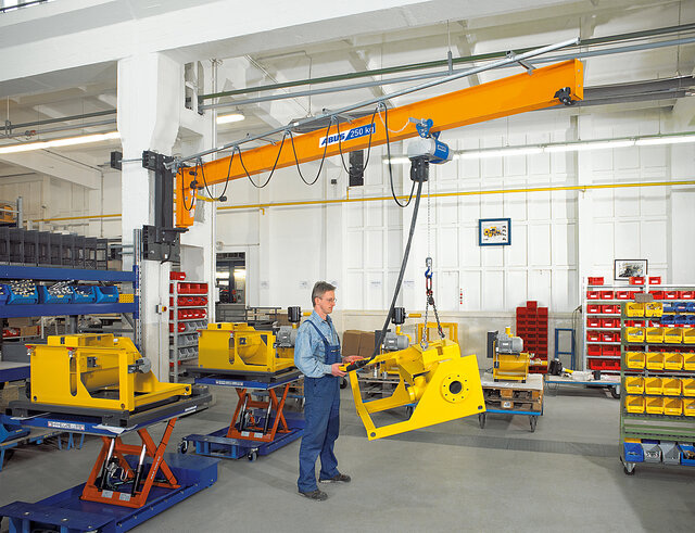 An employee operates a wall-mounted jib crane and moves a load using a chain hoist in a factory hall