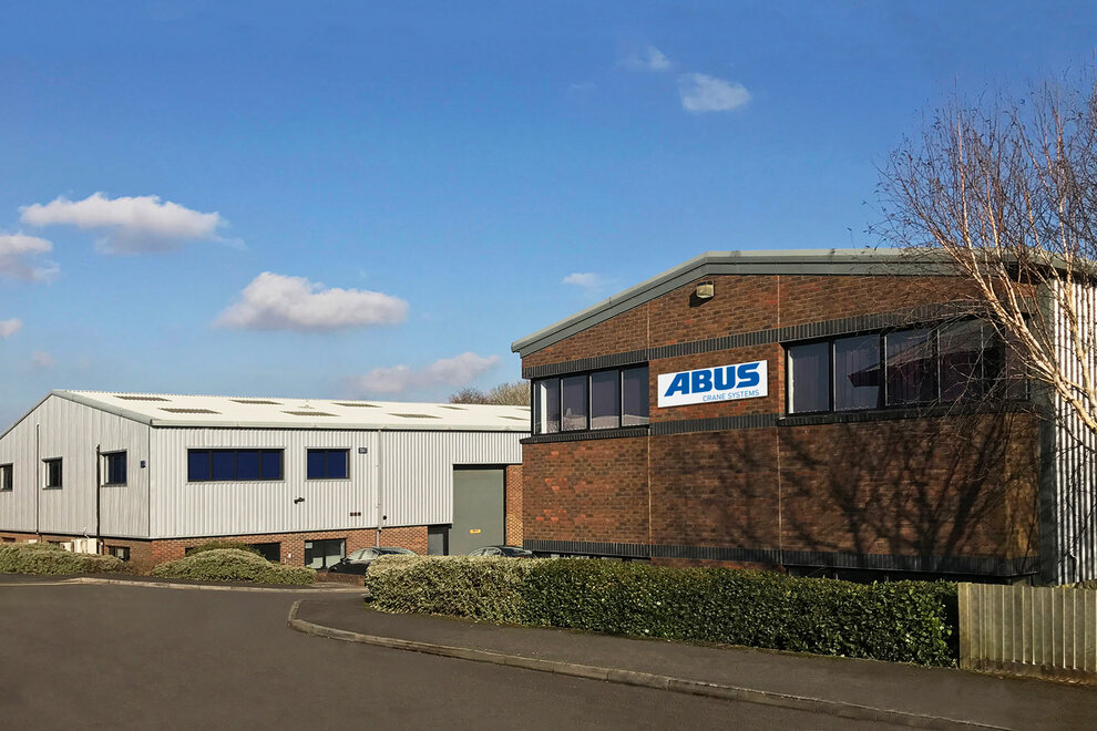 View of an ABUS Crane Systems company building in the Great Britain with a brick facade and an adjacent modern metal production hall, surrounded by green hedges and an asphalt access road