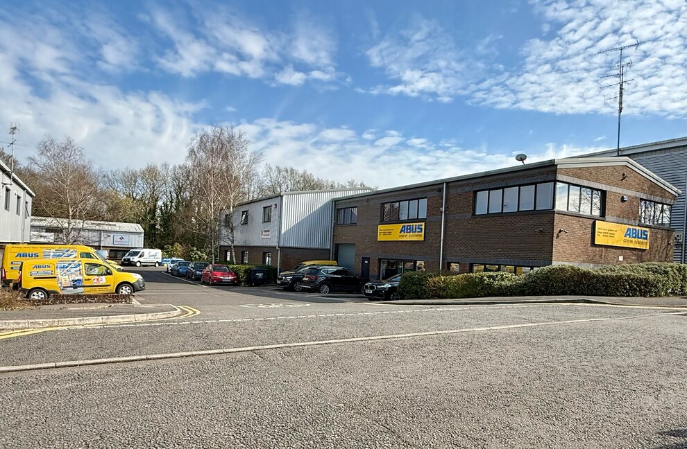 View of an ABUS Crane Systems company building in the Great Britain with a brick facade and an adjacent modern metal production hall, surrounded by green hedges and an asphalt access road