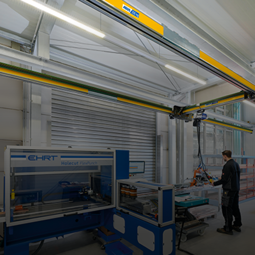 A jacked up single-girder crane EHB-X moves a load in an industrial hall, whilst an employee works on a machine in the background.